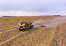 A yellow TerraGator sprayer equipped with booms is applying product across a large, open field under an overcast sky, with wind turbines visible in the distance.