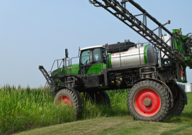 A Rogator sprayer is positioned near a field of tall corn, ready to apply liquid fertilizer under a clear blue sky.